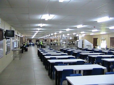 Long rows of tables in a large camp dining hall, with workers in hi-vis at the serving line